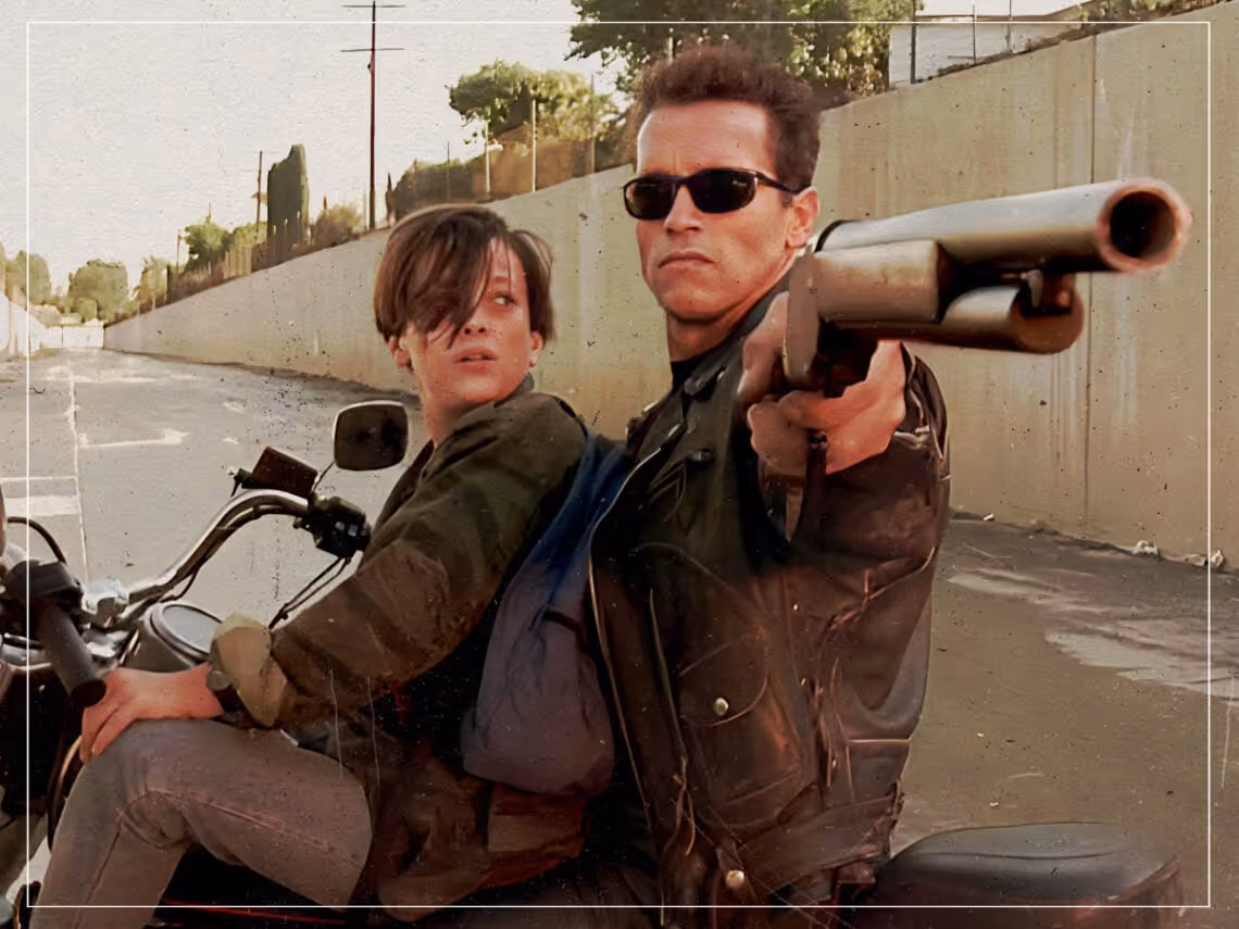 Two people on a motorcycle, a man wearing sunglasses and a leather jacket pointing a shotgun forward, with a woman sitting behind him looking backward.